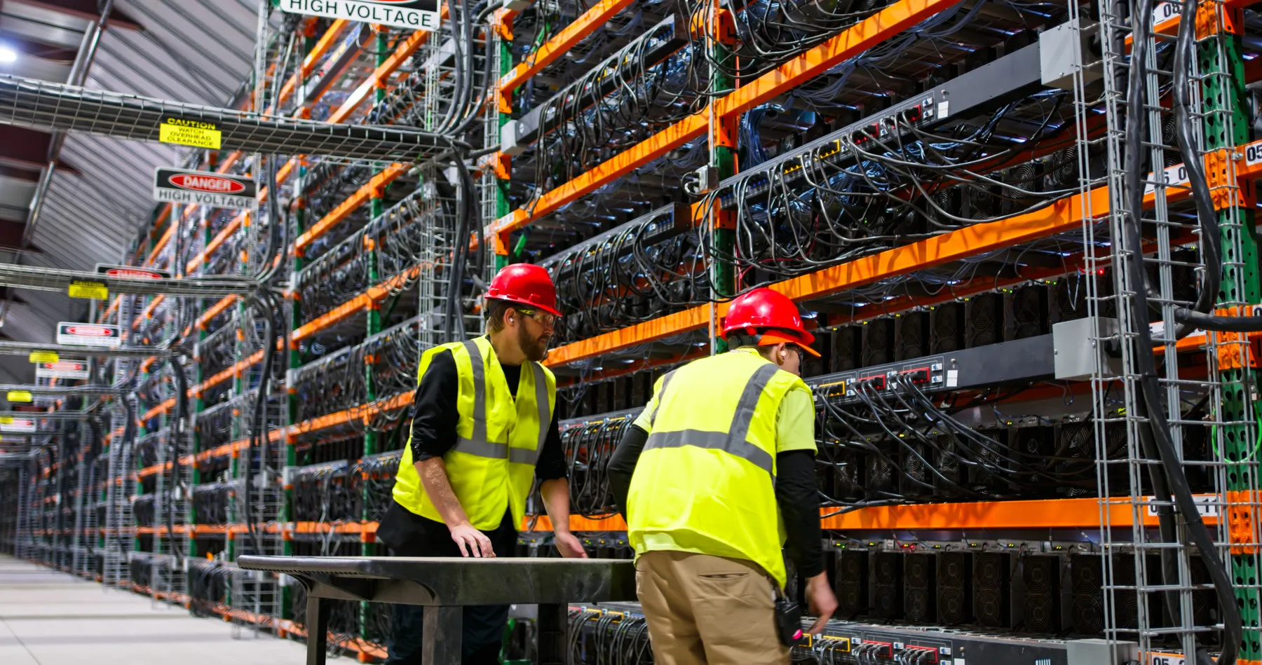 Two technicians wearing yellow vests walking past racks of electronic equipment in a data center for cryptocurrency mining.