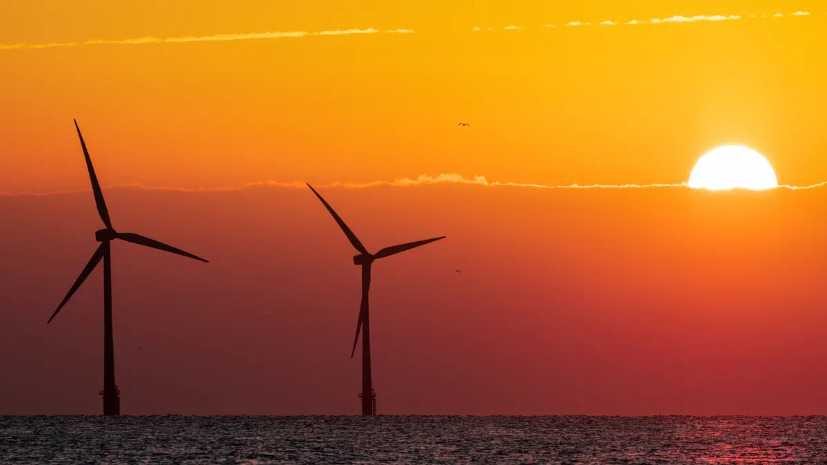 Offshore wind farm turbines against a graduated red sunset with the sun reflecting on the sea's horizon.